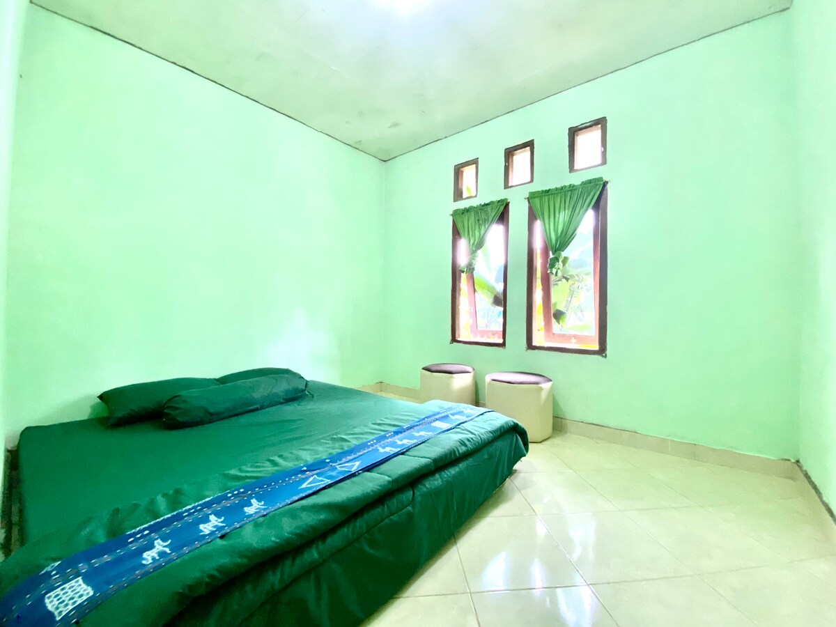 A spacious sleeping area features a large green bedspread laid over the floor, complemented by light green walls. Two windows allow natural light to fill the room, with green curtains framing the views. Two small ottomans are positioned nearby.