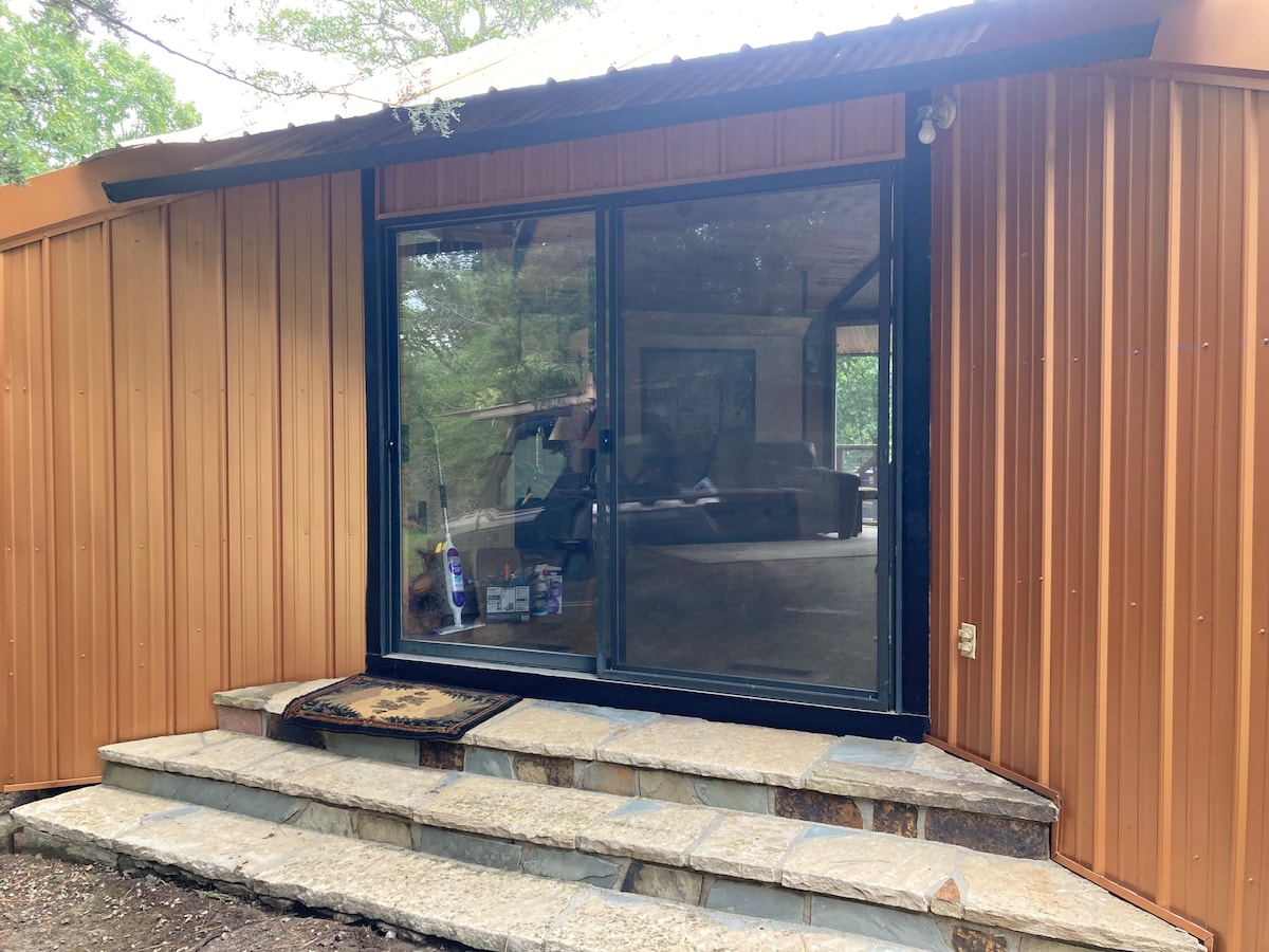 The entrance features a sliding glass door set within a warm-toned metal exterior. Stone steps lead to the doorway, providing access to the open living area visible beyond. Natural light is implied through the glass, blending outdoor and indoor spaces.
