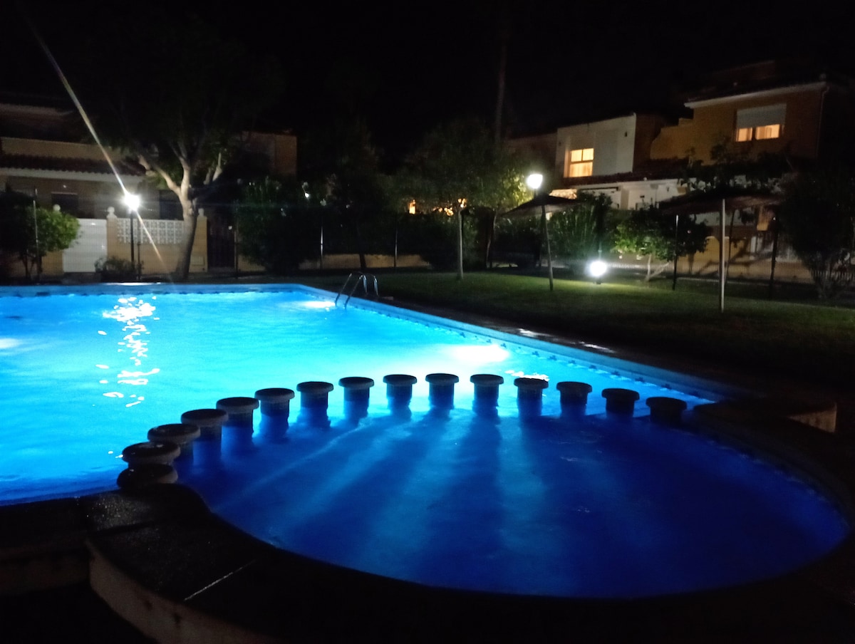 An illuminated swimming pool is featured at night, showcasing vibrant blue water. Surrounding greenery and subtle lighting enhance the serene atmosphere. A winding edge with circular seating is visible at the pool's side, providing a unique architectural element.