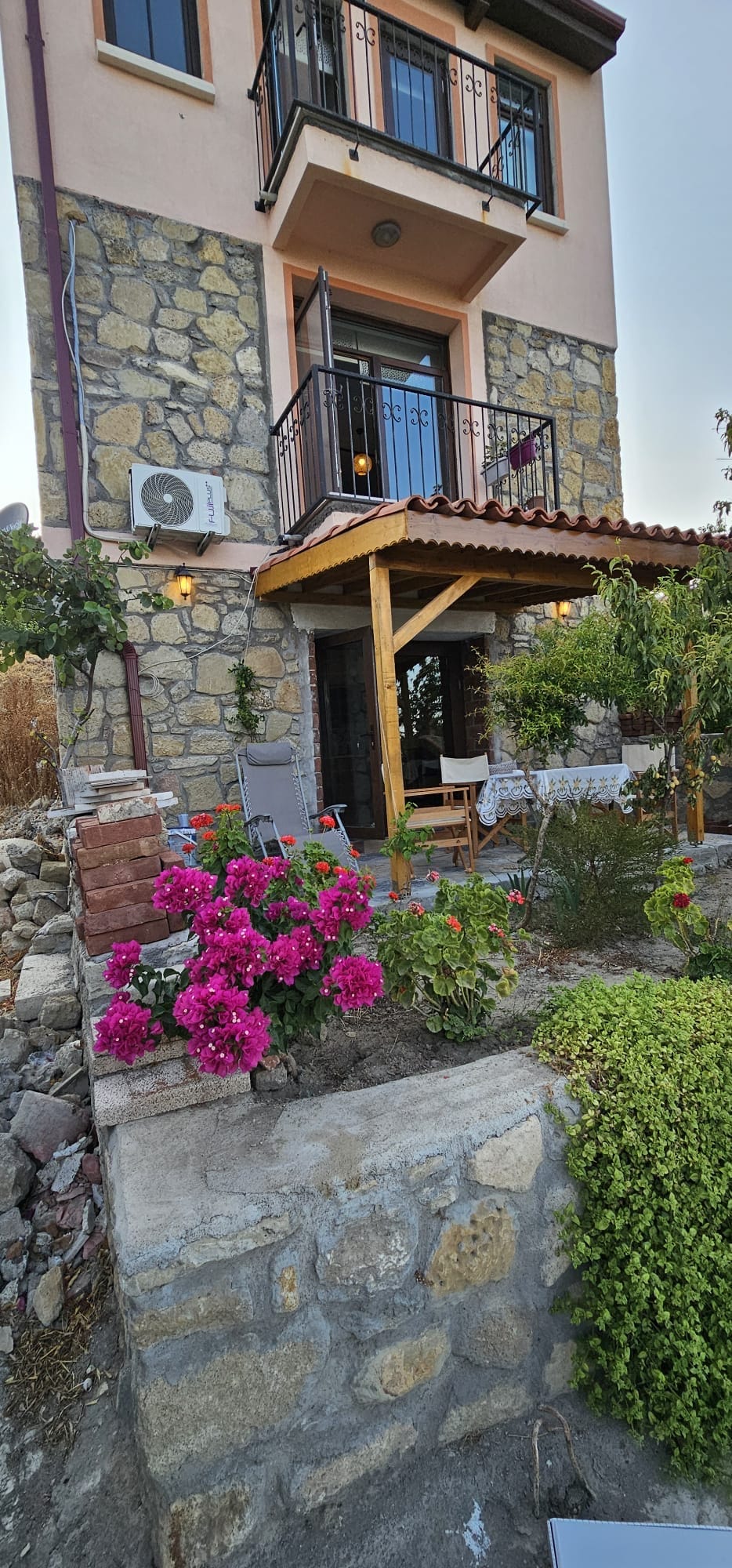 The exterior of a three-story stone house is prominently featured, with a wooden porch adorned with seating. Vibrant pink flowers and lush greenery enhance the entrance, creating a welcoming outdoor space. An air conditioning unit is positioned on the wall beside the porch.