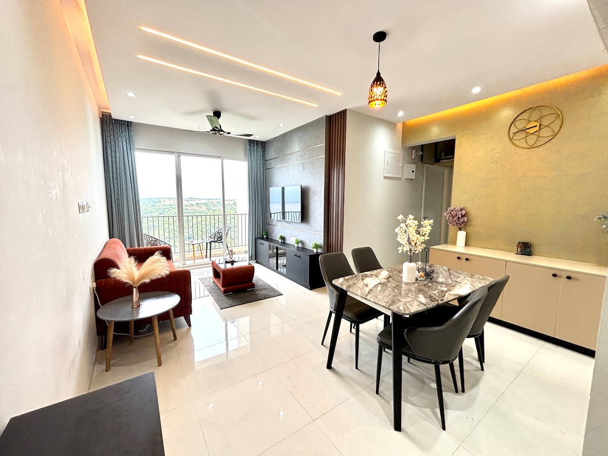 The living area features a modern design with polished white tiles and soft lighting. A cozy seating arrangement includes a pair of armchairs and a plush sofa facing a mounted TV. A dining table with six chairs is positioned next to a large window, showcasing a balcony view.