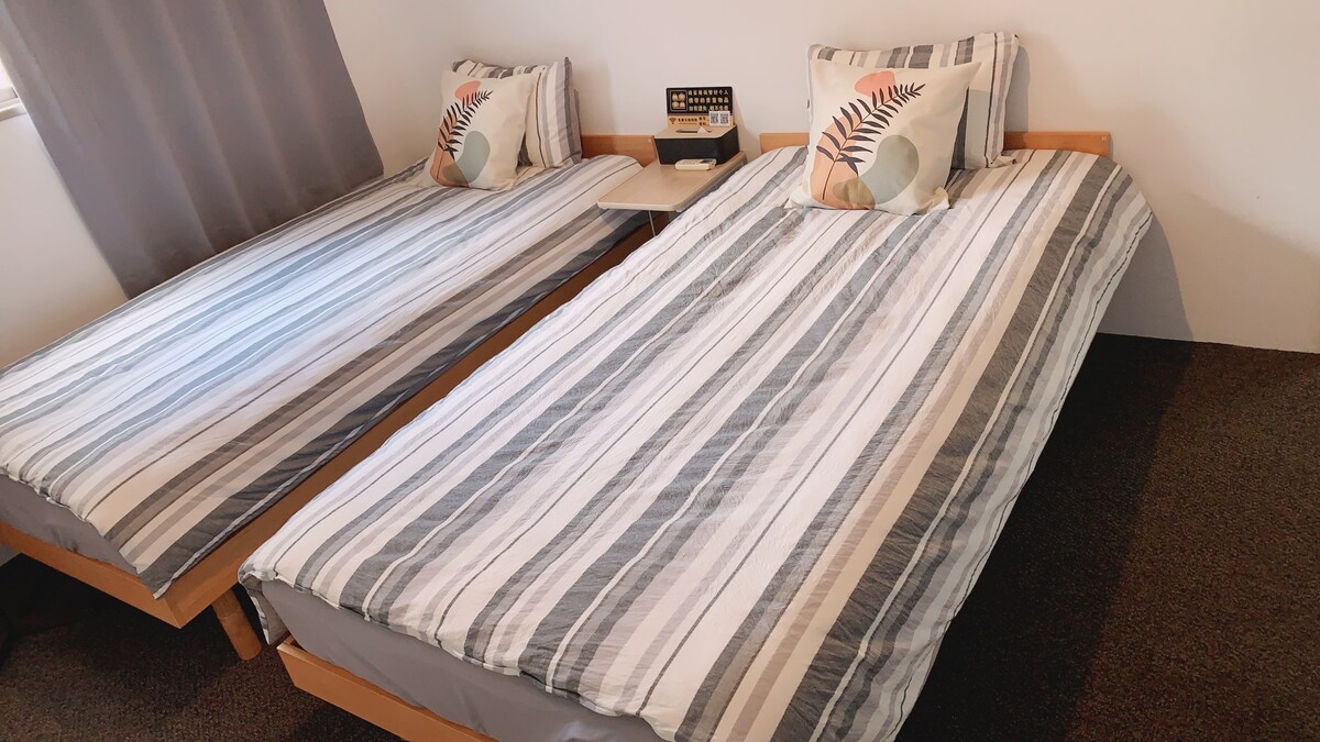 The room features two spacious beds, each adorned with striped bedding and decorative pillows. A bedside table is positioned between the beds, and natural light is noted filtering through a window with light gray curtains.
