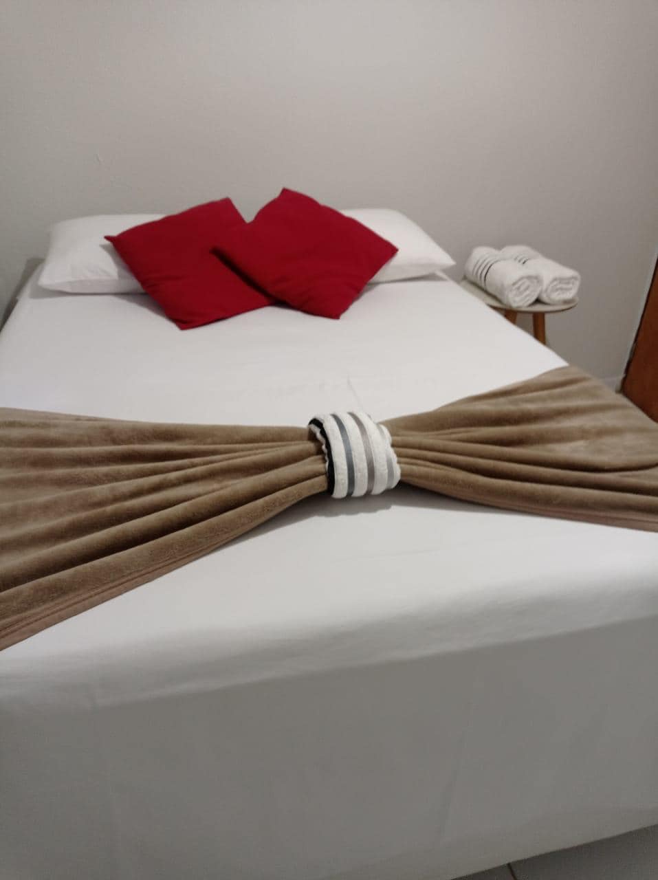 A comfortable bed with a light-colored sheet is centered in the room. Two red decorative pillows are placed at the head of the bed. A neutral-colored throw blanket is elegantly arranged at the foot, complemented by neatly rolled towels on the side.