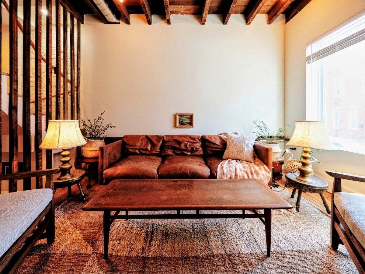 A comfortable living area features a worn-in leather sofa, complemented by a rustic coffee table and vintage lamps on either side. Natural light fills the space from the large window, illuminating the wooden ceiling beams and adding a warm touch to the neutral decor.