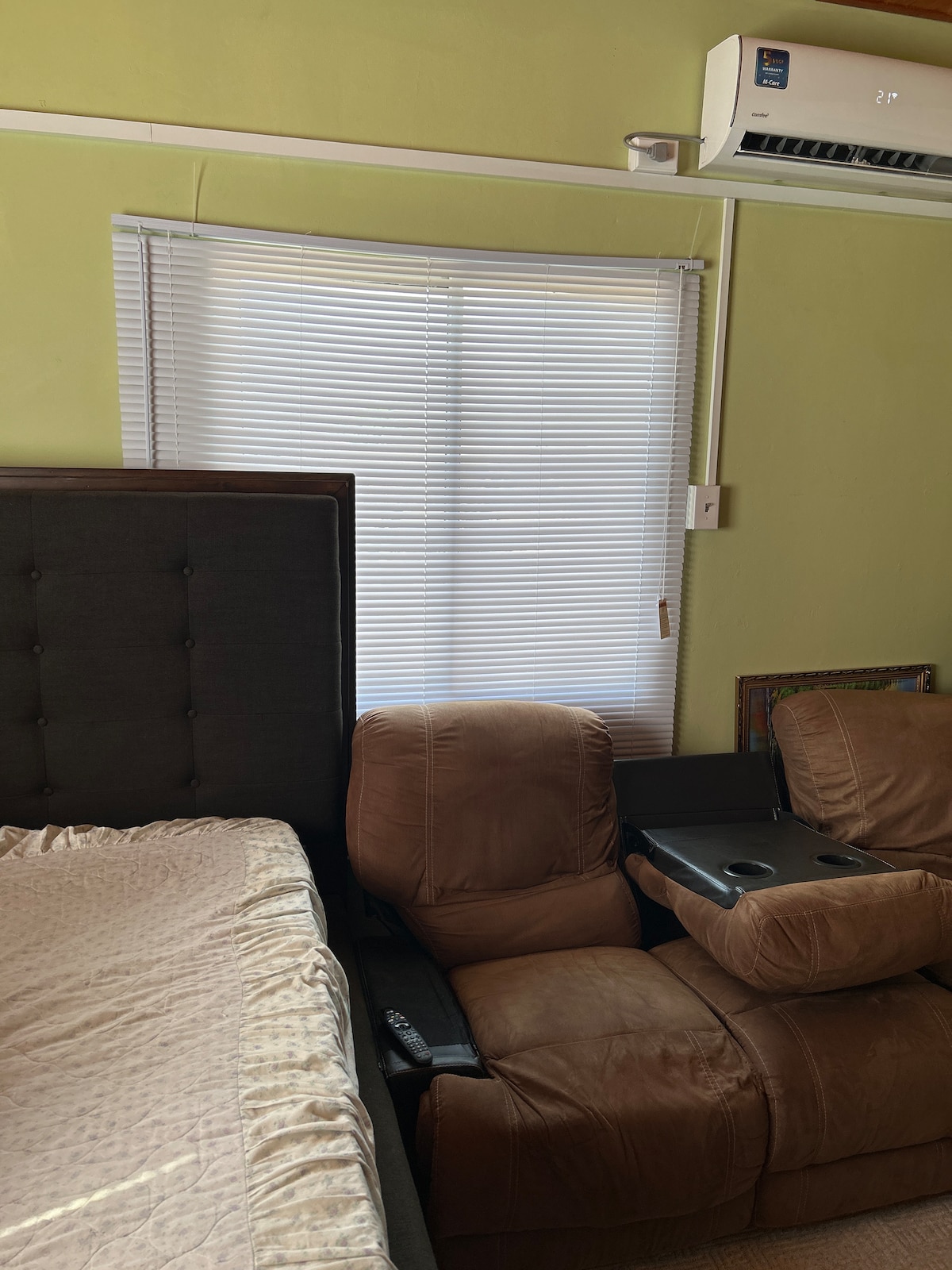 A compact sleeping area features a spacious bed with a dark headboard, complemented by a brown reclining chair equipped with built-in cup holders. Natural light filters in through a white window blind, enhancing the room's comfortable setting.