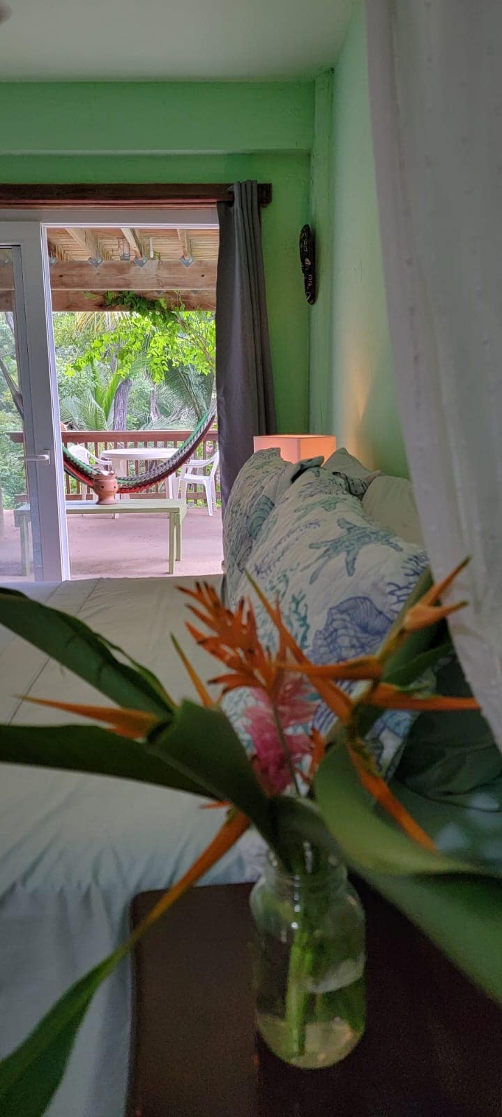 A spacious studio suite features soft green walls and inviting textiles. A cozy bed is adorned with decorative pillows. Outside, a large patio garden area is visible, with a hammock for relaxation. Tropical plants add vibrant color to the environment, enhancing the natural feel.