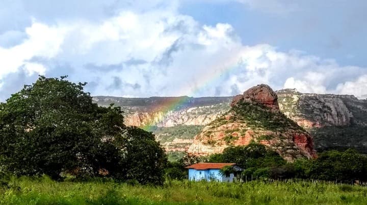 A scenic view features rolling hills and a vibrant rainbow arching across a cloudy sky. Lush greenery surrounds a small blue house nestled at the base of a rocky outcrop, highlighting the natural beauty of the landscape.