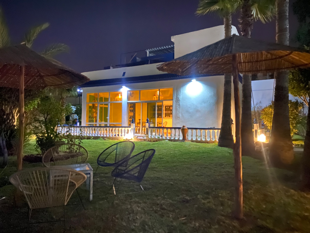 The villa is illuminated at night, showcasing large glass windows that reveal a welcoming interior. A spacious lawn is equipped with seating areas under thatched umbrellas. Lush greenery and palm trees surround the outdoor space, creating a serene atmosphere.