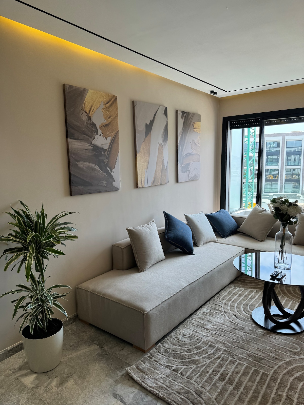 A modern living area features a comfortable sectional couch adorned with cushions in varied shades of blue and gray. Above, three abstract paintings provide a subtle artistic touch. A small round table sits on a patterned rug, complemented by a potted plant in the corner.