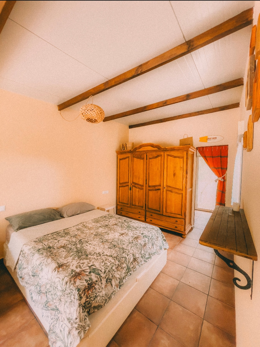 A welcoming bedroom features a spacious bed covered with a patterned blanket. Natural light enters through the window, which is adorned with a light curtain. A traditional wardrobe made of wood stands against the wall, while a simple wooden shelf is positioned beside the bed.