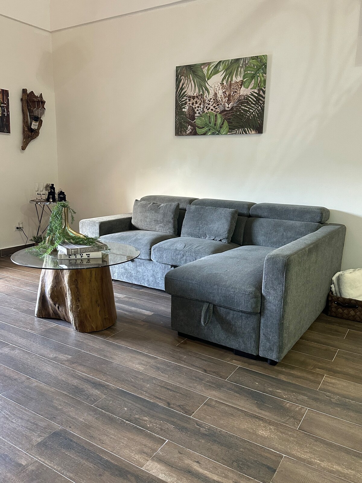 A gray sectional sofa is positioned beside a small, round glass table with a natural wood base. A piece of tropical wall art hangs above the sofa, enhancing the room's relaxed ambiance. The flooring features warm-toned wooden planks, creating a cozy living area.