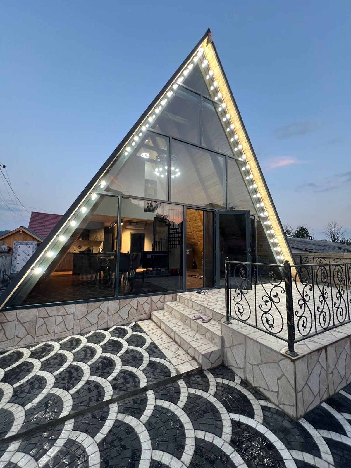 An A-frame structure features large glass panels, allowing natural light to fill the interior. The entrance is framed by a patterned stone pathway, leading up to a set of stairs. Decorative lighting outlines the roof, enhancing the architectural design.