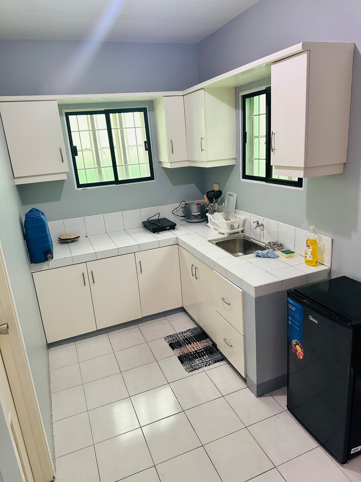 A well-equipped kitchenette features light-colored cabinets and a clean countertop. Natural light enters through two windows, illuminating a stainless steel sink and various appliances. A small refrigerator is situated in the corner, with a black mat on the tiled floor.