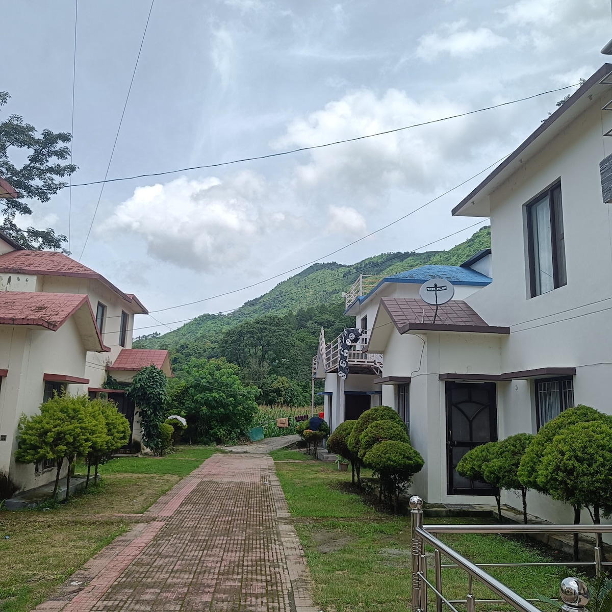 A peaceful pathway is lined with two-story homes, each featuring a pitched roof and well-maintained gardens. Green, manicured shrubs frame the walkway, while a lush mountain backdrop rises dramatically in the distance under a sky scattered with soft clouds.