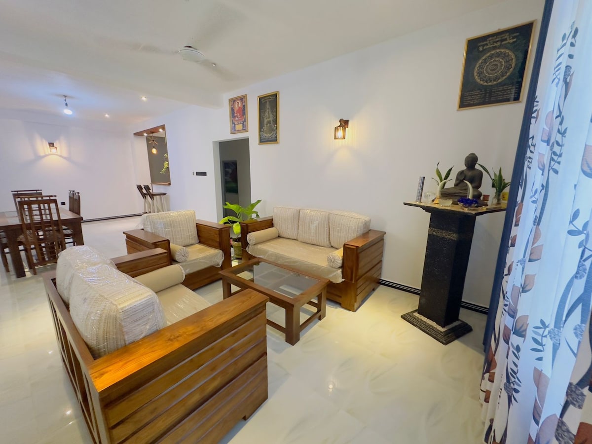 A spacious living area features two wooden couches with light-colored cushions positioned around a low coffee table. A decorative plant is placed nearby. The room is well-lit, with soft lighting fixtures and a wall adorned with artwork, creating a calm atmosphere.