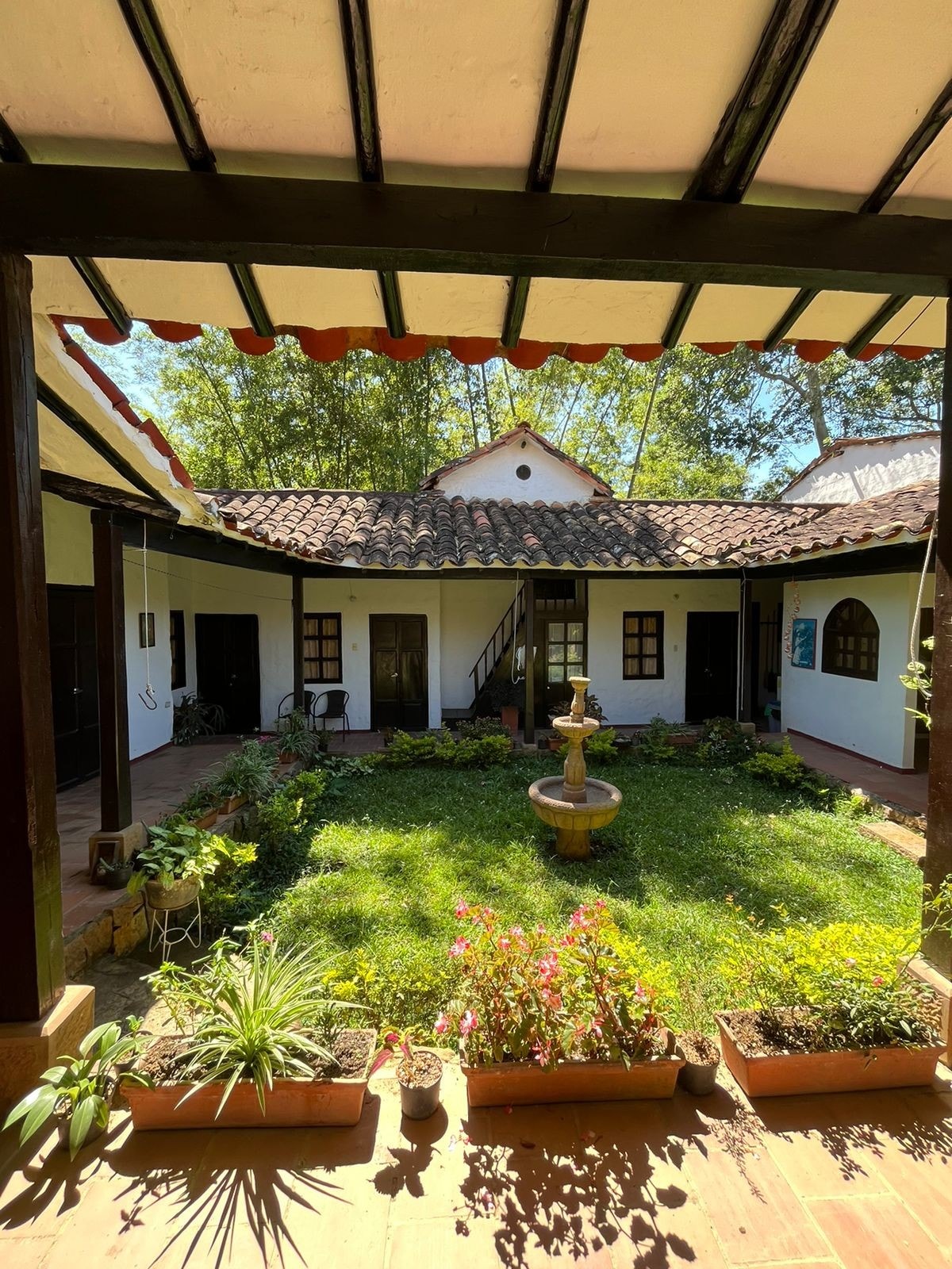 A courtyard is encompassed by colonial-style architecture, featuring a central fountain surrounded by lush greenery and blooming flowers. The structure showcases wooden beams and arched windows, creating a peaceful outdoor space that invites relaxation and connection.