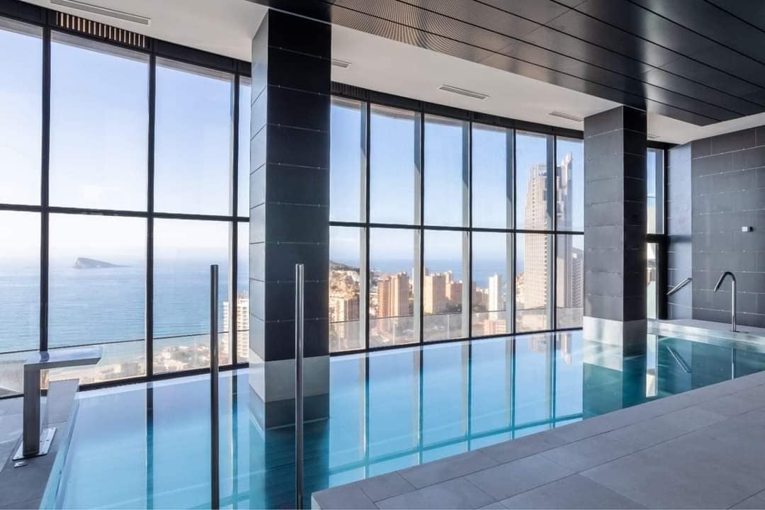 An indoor pool area is presented, featuring large floor-to-ceiling windows that provide expansive views of the sea and surrounding city. The water reflects light, and sleek gray tiles adorn the floor, enhancing the contemporary aesthetic of the space.