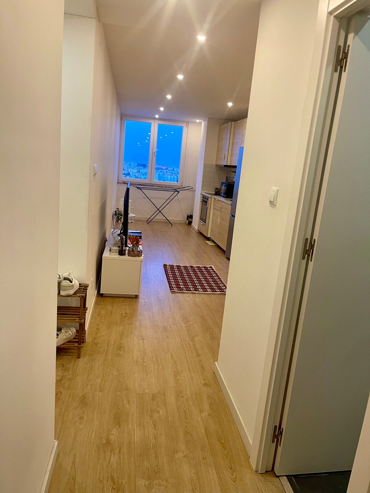 A long hallway leads to an open kitchen space, featuring warm wood flooring and warm lighting. The kitchen is equipped with modern appliances, and a small white table is placed near the window, offering a view of the city skyline.