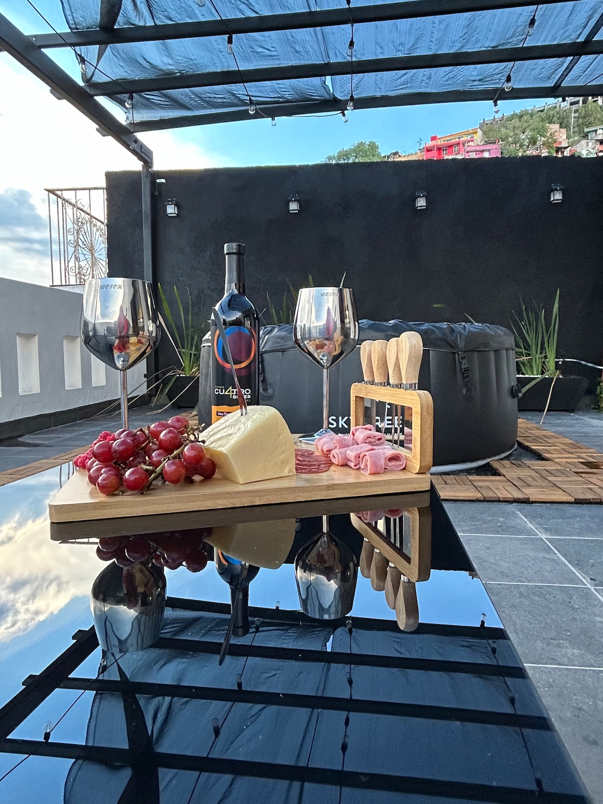 A stylish outdoor terrace is presented, featuring a black hot tub surrounded by plants. A polished surface holds a wooden cheese board with grapes, cheese, and cured meats, complemented by two glasses and utensils, all under a protective canopy.