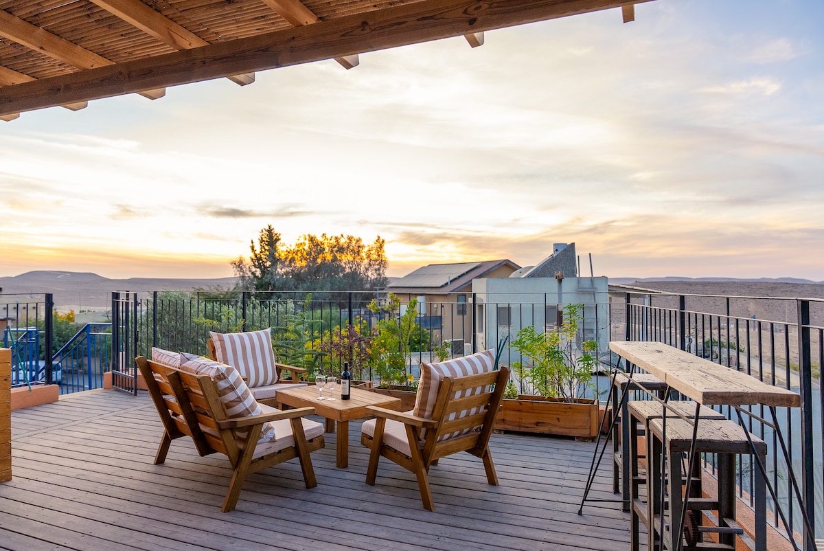 A spacious terrace is featured, showcasing wooden lounge chairs and a rustic table positioned for sunset views. Potted plants add greenery, and a high-top table stands nearby. A vast desert landscape is visible in the background, highlighted by a soft evening glow.