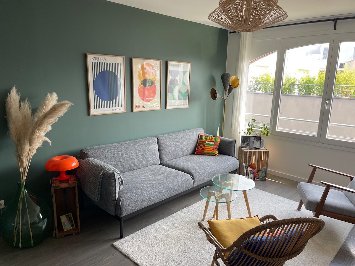 A cozy living area is illuminated by natural light from large windows. A grey sofa is complemented by colorful cushions and decorative artwork on a green wall. A round glass coffee table is surrounded by diverse seating options, including a wicker chair and a stylish lamp.