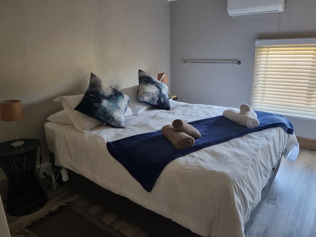 A well-lit bedroom features a spacious bed adorned with two patterned cushions and a dark throw blanket. Two rolled towels are neatly placed on the bed, while a bedside table with a lamp is positioned beside it. Natural light enters through a partially closed window.