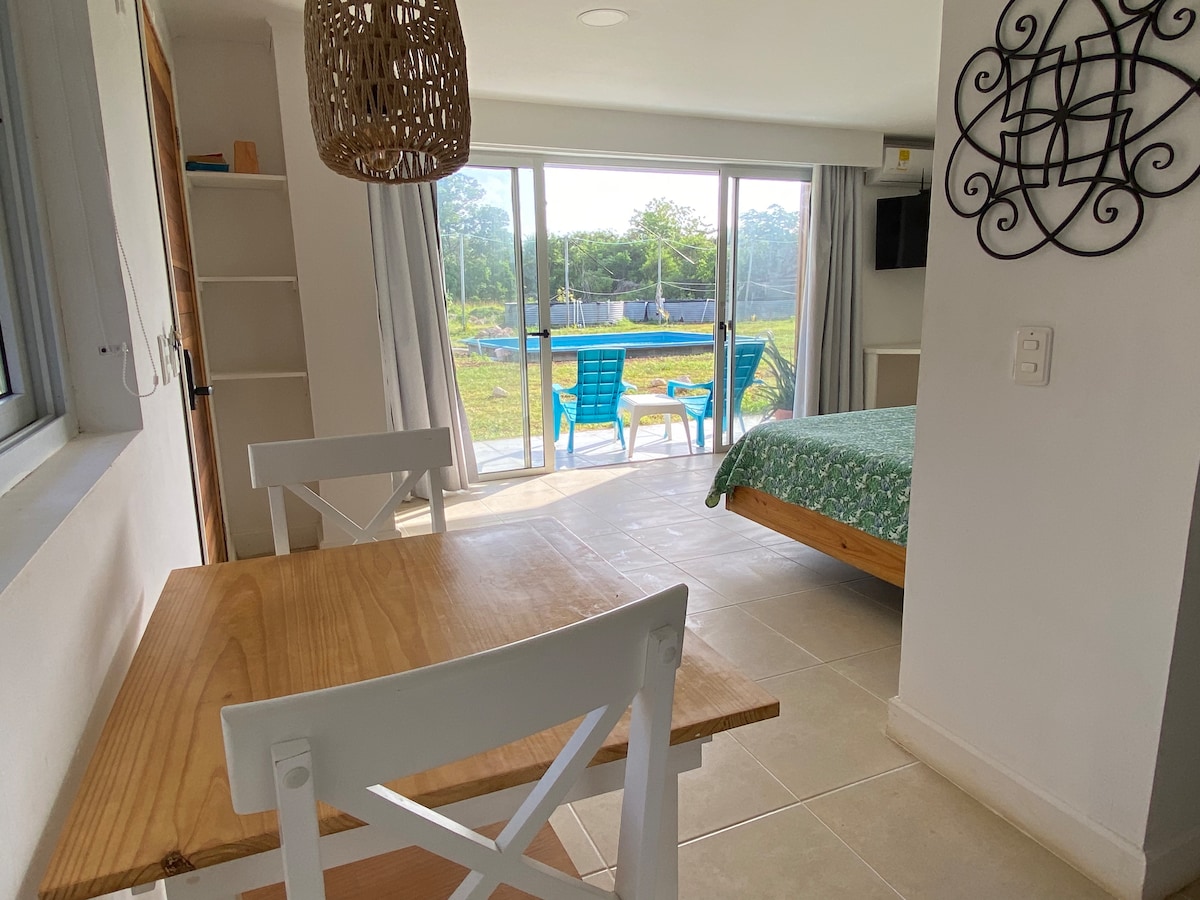 A dining area features a light wood table with two white chairs, positioned near a large sliding glass door. Beyond the door, a green landscape is visible, showcasing outdoor seating with blue chairs and a pool area, while a cozy sleeping space is highlighted by a green-patterned bed covering.