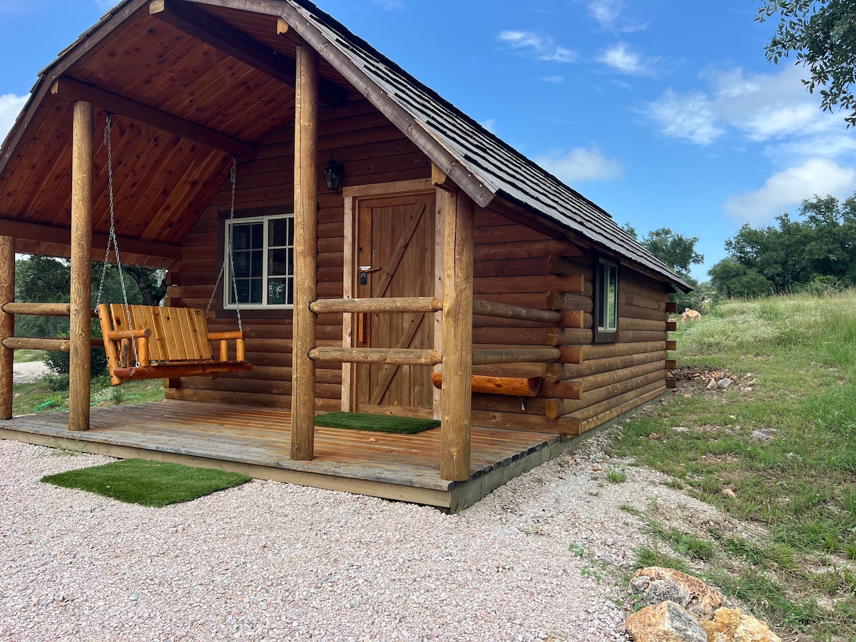 A rustic log cabin is presented with a covered porch featuring a wooden swing. The cabin's exterior showcases natural wood textures, complemented by a gravel area and green grass in the surrounding landscape. Blue skies and scattered clouds create a serene backdrop.