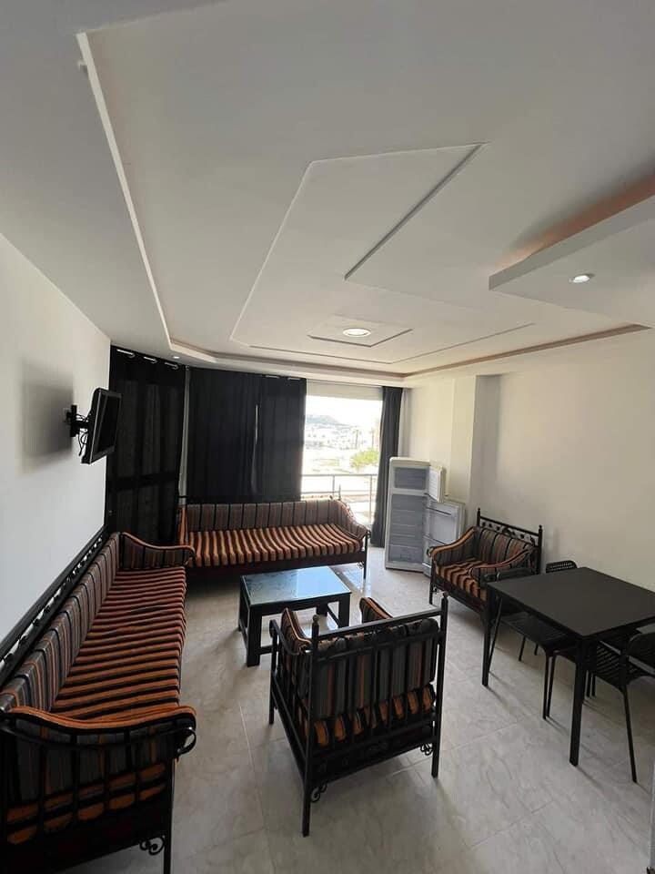A bright living area features multiple seating arrangements, including a striped sofa and several chairs around a black table. Large windows allow natural light to enter, while dark curtains provide optional privacy. A television is mounted on one wall.