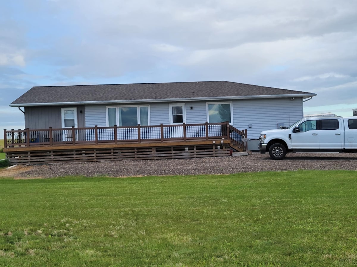 A single-story house is visible, featuring a broad wooden deck with railings. Large windows allow natural light to enter the interior. A truck is parked nearby on a gravel surface, with a green lawn extending in the foreground.