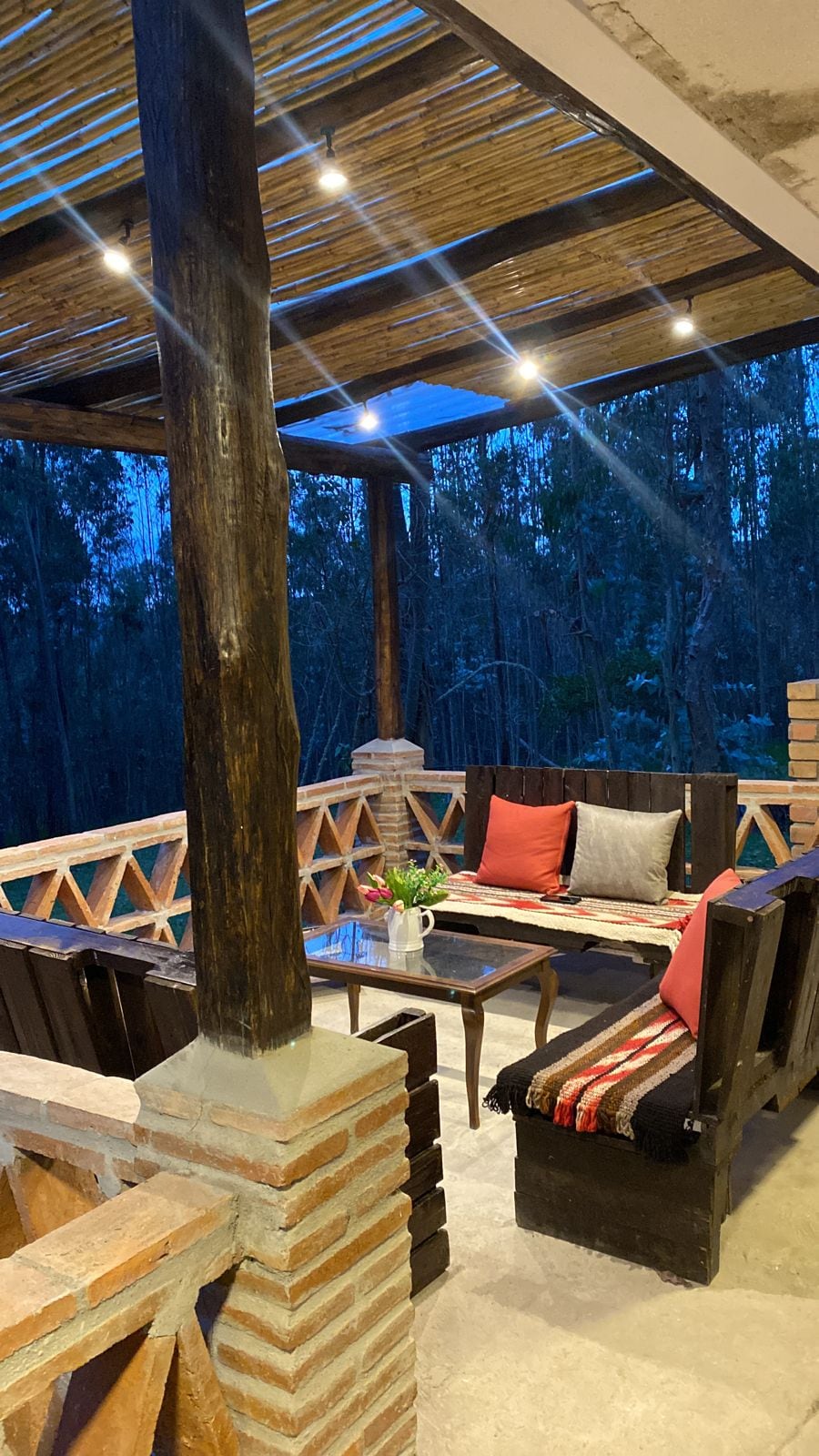 An outdoor seating area is presented under a wooden pergola, enhanced by soft lighting. The arrangement includes a glass table surrounded by chairs with cushions. Eucalyptus trees are visible in the background, contributing to a serene environment.