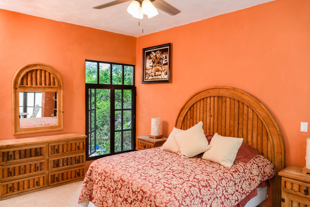The bright bedroom features an earthy orange wall, a spacious bed with patterned bedding, and two pillows. A large, framed artwork hangs above the bed. A wooden dresser and mirror are positioned against one wall, while a set of glass doors allows natural light and views of greenery.