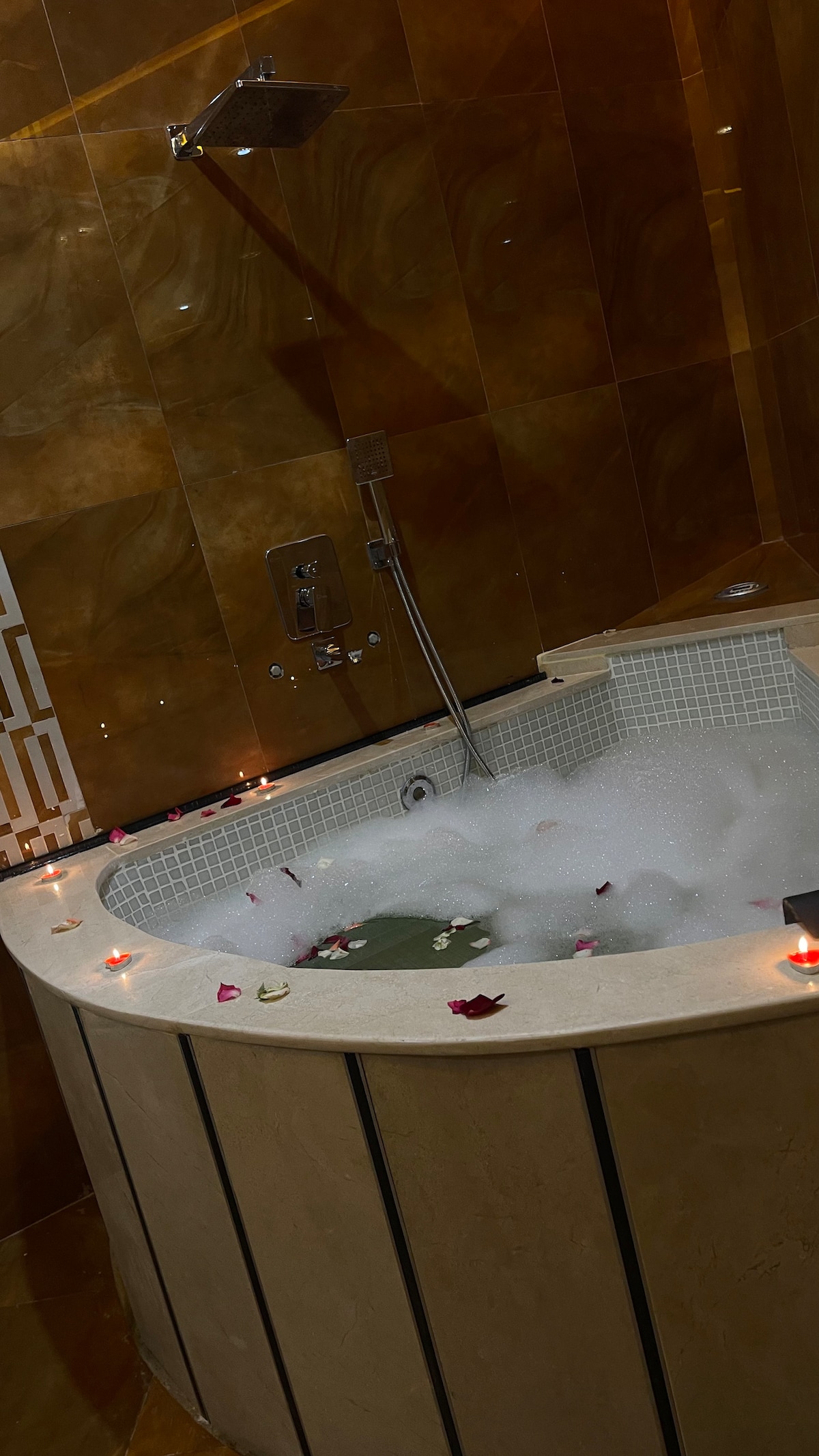 A circular jacuzzi is positioned within a marble surround, filled with white bubbles and accented by scattered rose petals. Soft candles provide a gentle glow, and warm, earthy tones of the tiles create a soothing backdrop for relaxation.