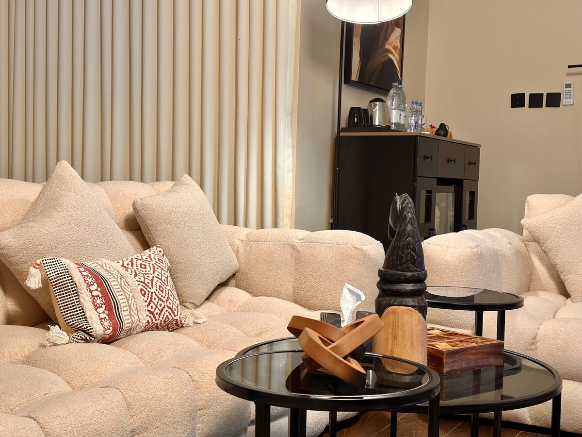 A comfortable seating area features a plush sectional sofa in a neutral tone, accented by decorative cushions. Nestled nearby are two sleek black coffee tables, showcasing a wooden tray and a carved sculpture. Soft lighting from a modern lamp creates a warm ambiance.
