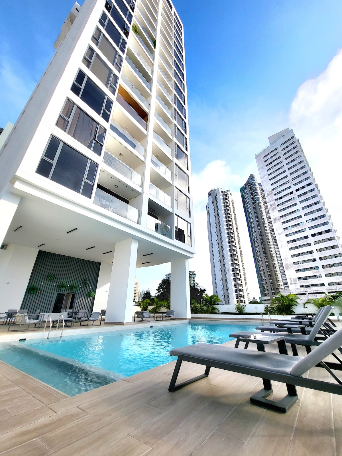 A modern pool area is highlighted with lounge chairs positioned around the water. The building's sleek exterior rises in the background, complemented by tall neighboring structures. The clear sky adds to the vibrant atmosphere of the space, inviting relaxation.