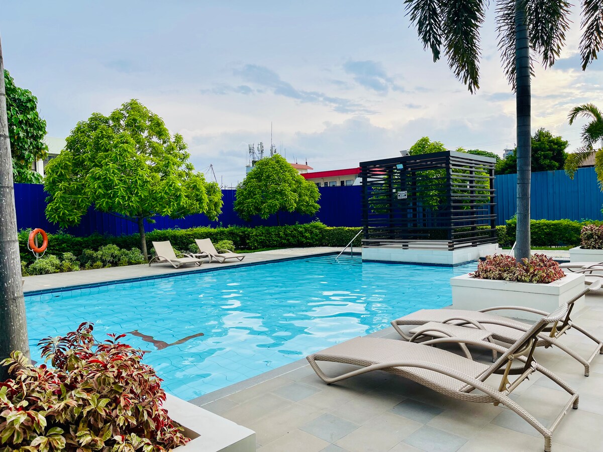 A shared pool is framed by tropical plants and features lounge chairs on a stone deck. A shaded seating area is present in the background, offering a place to relax. The serene water reflects the clear sky, creating a tranquil retreat.