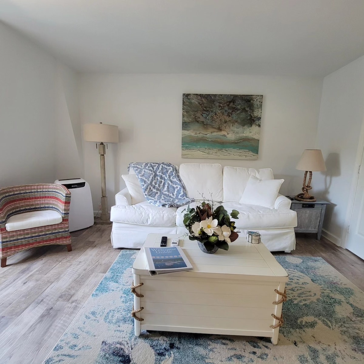 The living area features a white couch adorned with decorative pillows and a soft throw. A patterned armchair complements the space. A neutral-colored coffee table sits at the center, surrounded by a light area rug. Soft lighting is provided by lamps on either side.