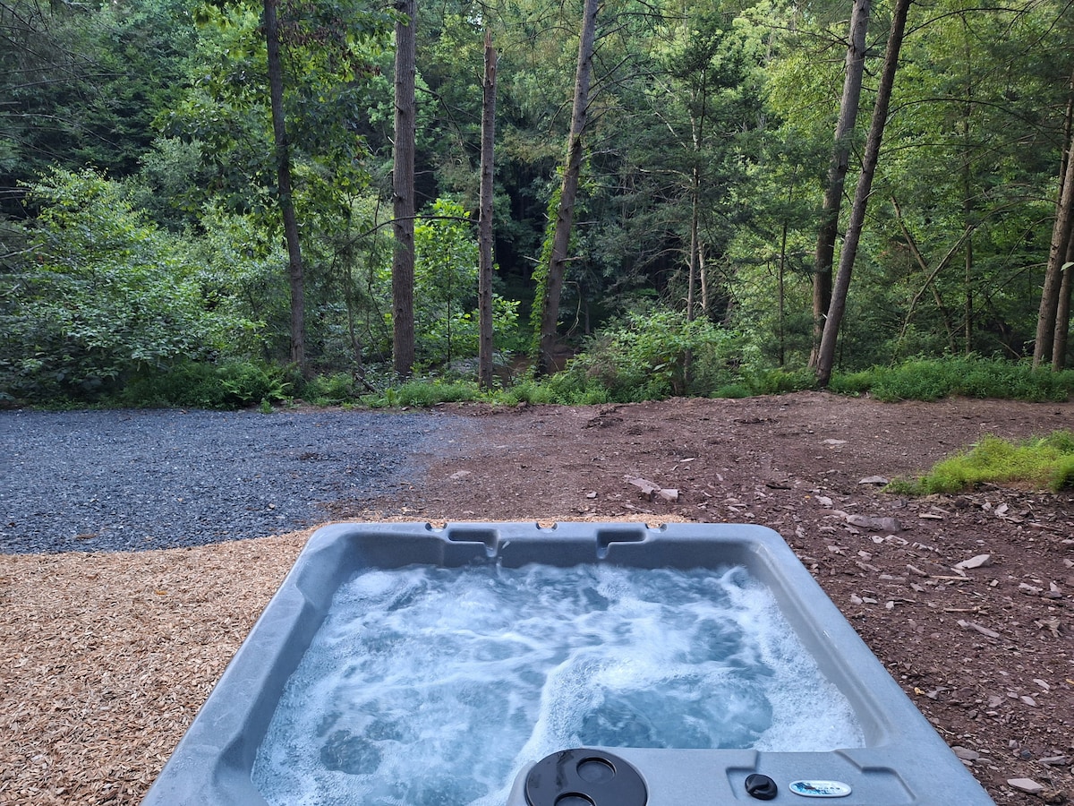 A hot tub sits on a gravel area, surrounded by trees that lend a sense of privacy. The bubbling water invites relaxation with a natural backdrop of forest greenery. The scene conveys a peaceful outdoor retreat, ideal for enjoying nature.
