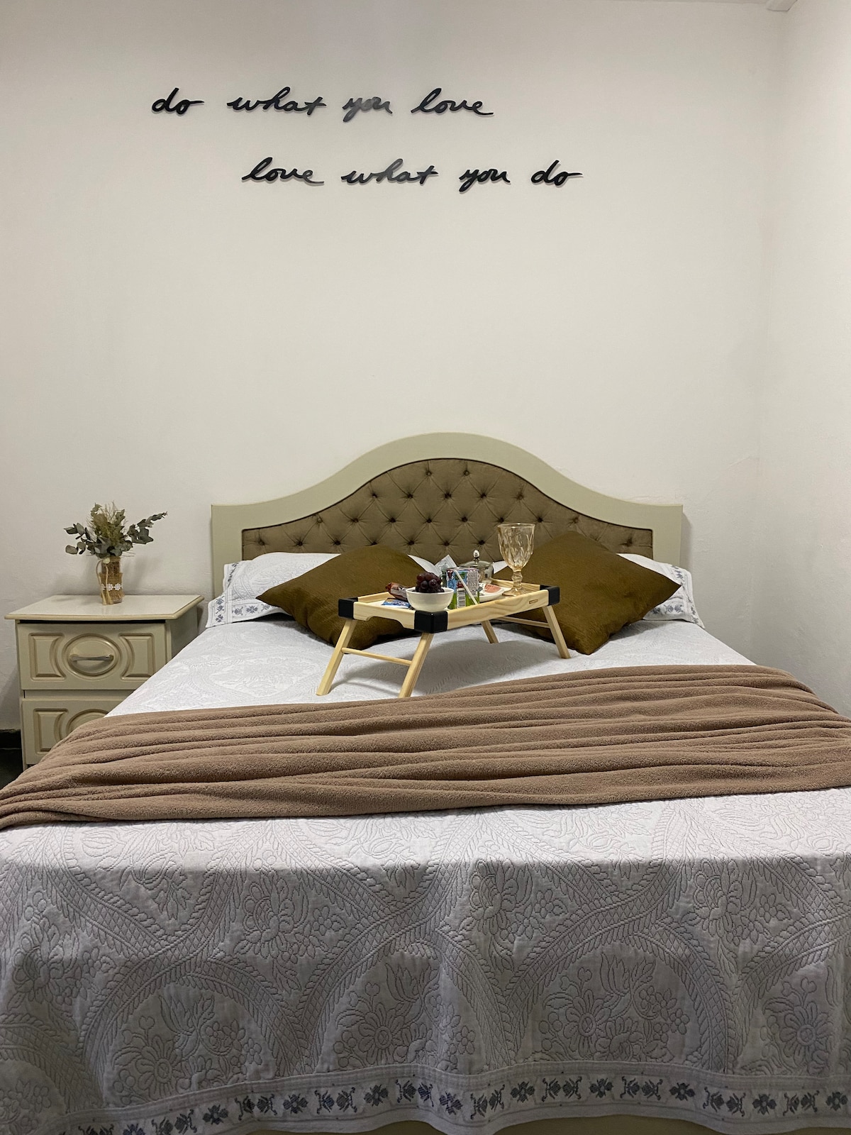 A comfortable sleeping area features a spacious bed with a decorative headboard. Soft brown pillows accent the bed, which is layered with a light blanket. A bedside table holds a small tray with beverages and snacks. A motivational phrase is displayed on the wall above the bed.