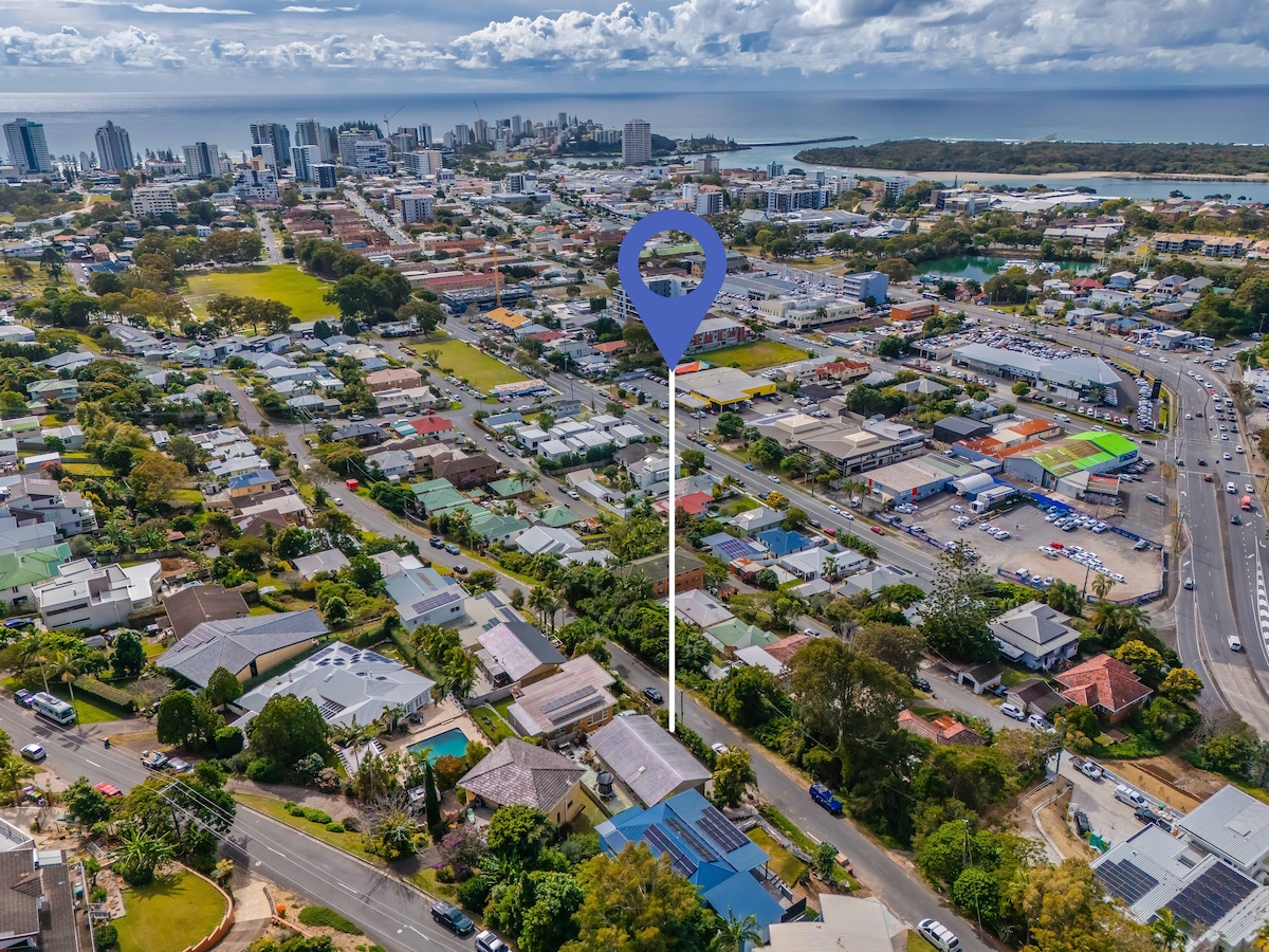 An aerial view captures the surrounding area of Tweed Heads, featuring a mix of residential homes, green spaces, and commercial buildings. The coastline and distant city skyline are visible, with roads and vehicles creating a sense of accessibility to nearby attractions.