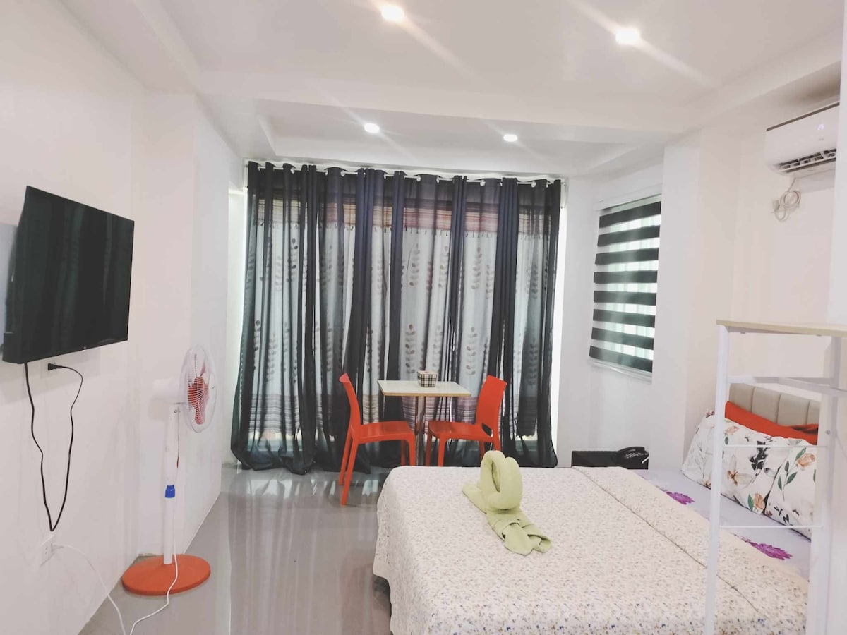 A spacious room is highlighted by a large window adorned with dark curtains, allowing natural light to fill the space. A comfortable bed is accompanied by decorative pillows and a folded towel. A small dining table with orange chairs and a wall-mounted television are also visible.