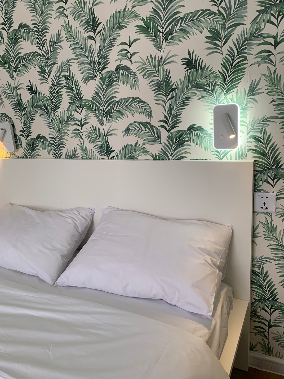 A spacious bed is positioned against a wallpaper adorned with lush green palm leaves. Soft, white bedding and two plush pillows provide comfort. Wall-mounted lamps are placed on either side, offering ambient light for reading.