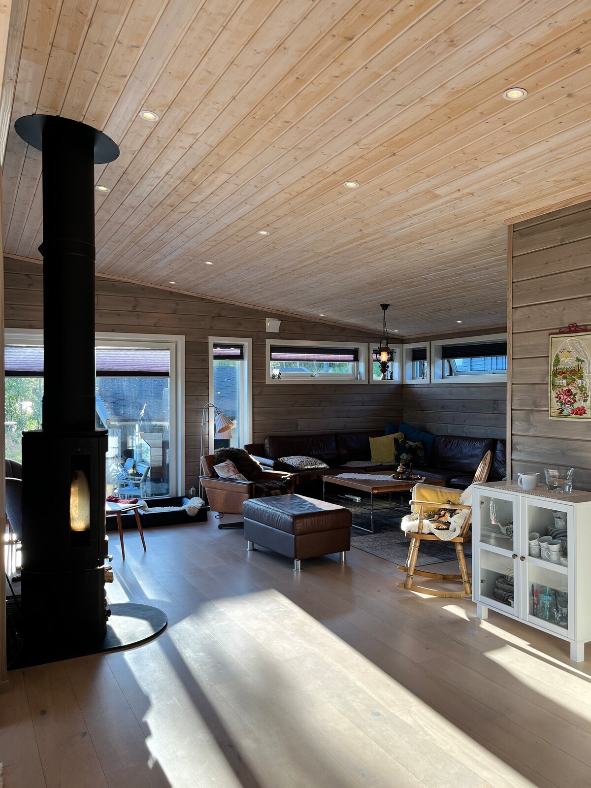 A spacious living area is seen with large windows allowing natural light to illuminate the wood-paneled walls. A cozy seating arrangement includes a sectional sofa and leather chairs, positioned around a central coffee table. A modern stove stands on one side, contributing to the cabin's warmth.