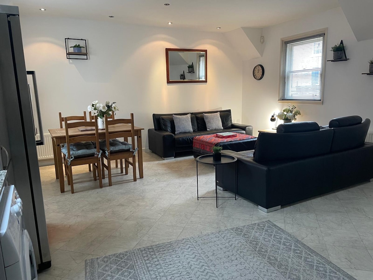 An open plan kitchen and living area is featured, containing a dining table with wooden chairs and a spacious sofa. A decorative mirror hangs on the wall, while light enters through windows adorned with simple treatments. A soft area rug adds texture to the floor.