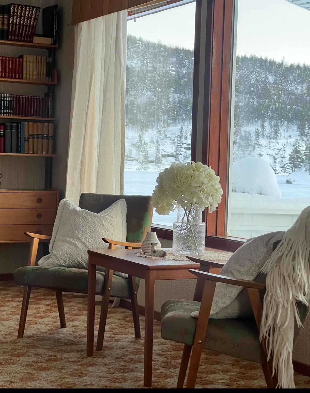 Two armchairs are positioned next to a small wooden table, creating a cozy seating area. A vase filled with white flowers sits atop the table. Large windows offer views of snow-covered hills and a winter landscape, allowing natural light to fill the space.