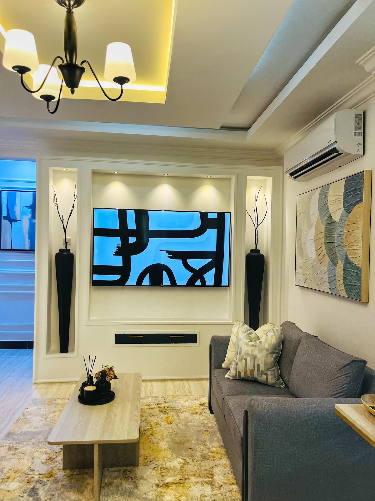 A modern living area is presented with a gray sofa and a stylish coffee table. Ambient lighting highlights the artwork and two tall decorative vases. A flat-screen television is mounted on the wall, framed by a light-colored panel, creating a contemporary focal point.
