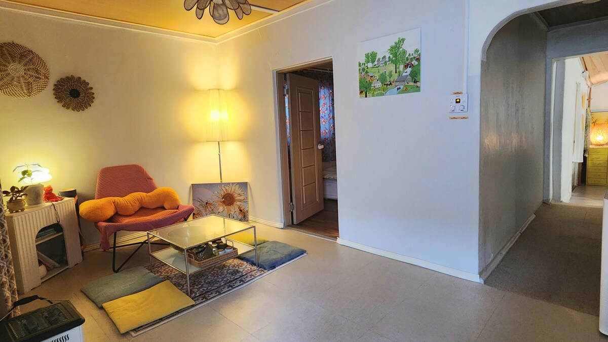 A functional living space is shown, featuring a glass coffee table set on a patterned rug. Soft cushions are placed on the floor, while a cozy chair highlights the corner. Natural light enters through a partially curtained window, illuminating the neutral-toned walls.