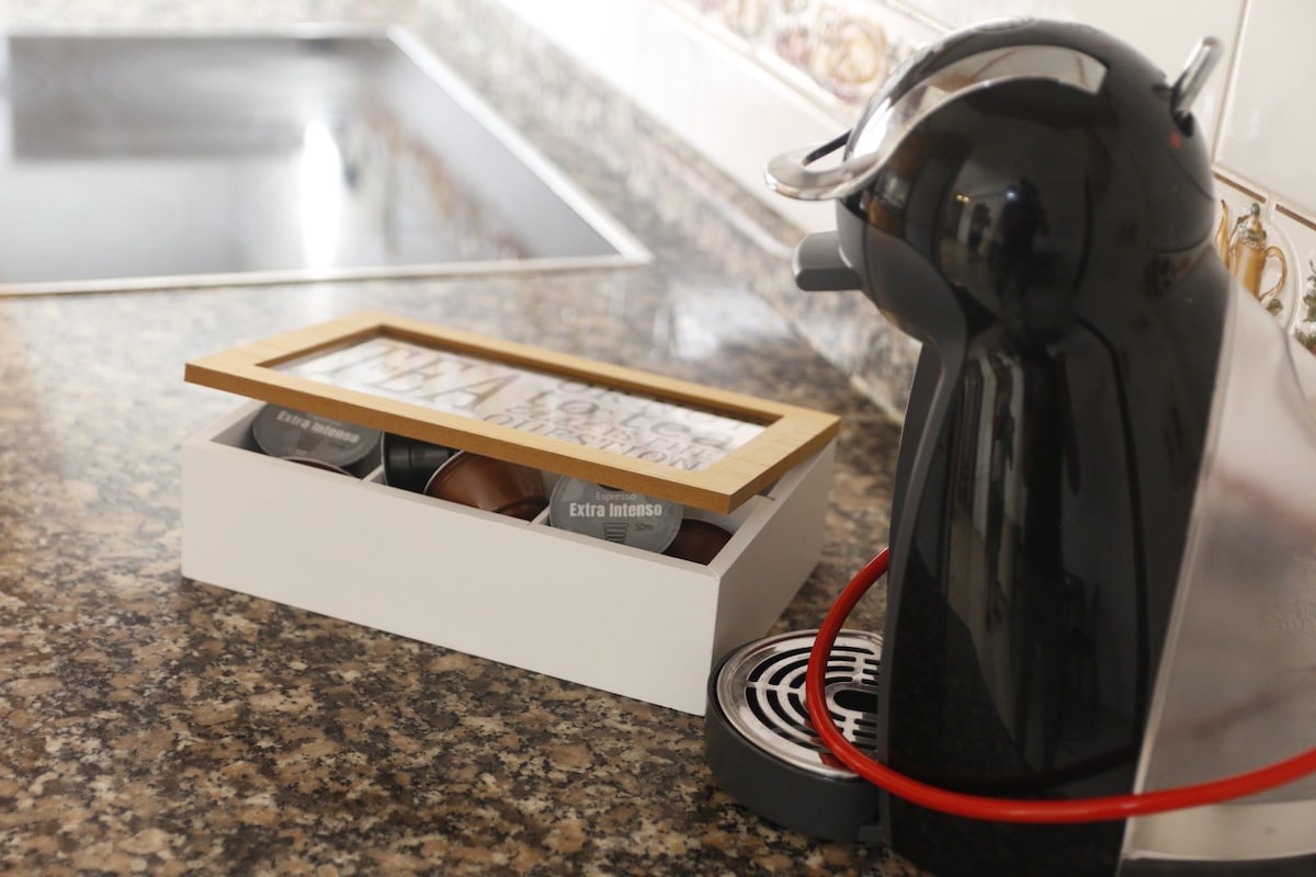 A modern coffee machine is positioned next to a sleek countertop. A white box contains various coffee capsules, topped with a wooden frame displaying a decorative image. The textured surface of the granite counter contrasts with the glossy finish of the machine.