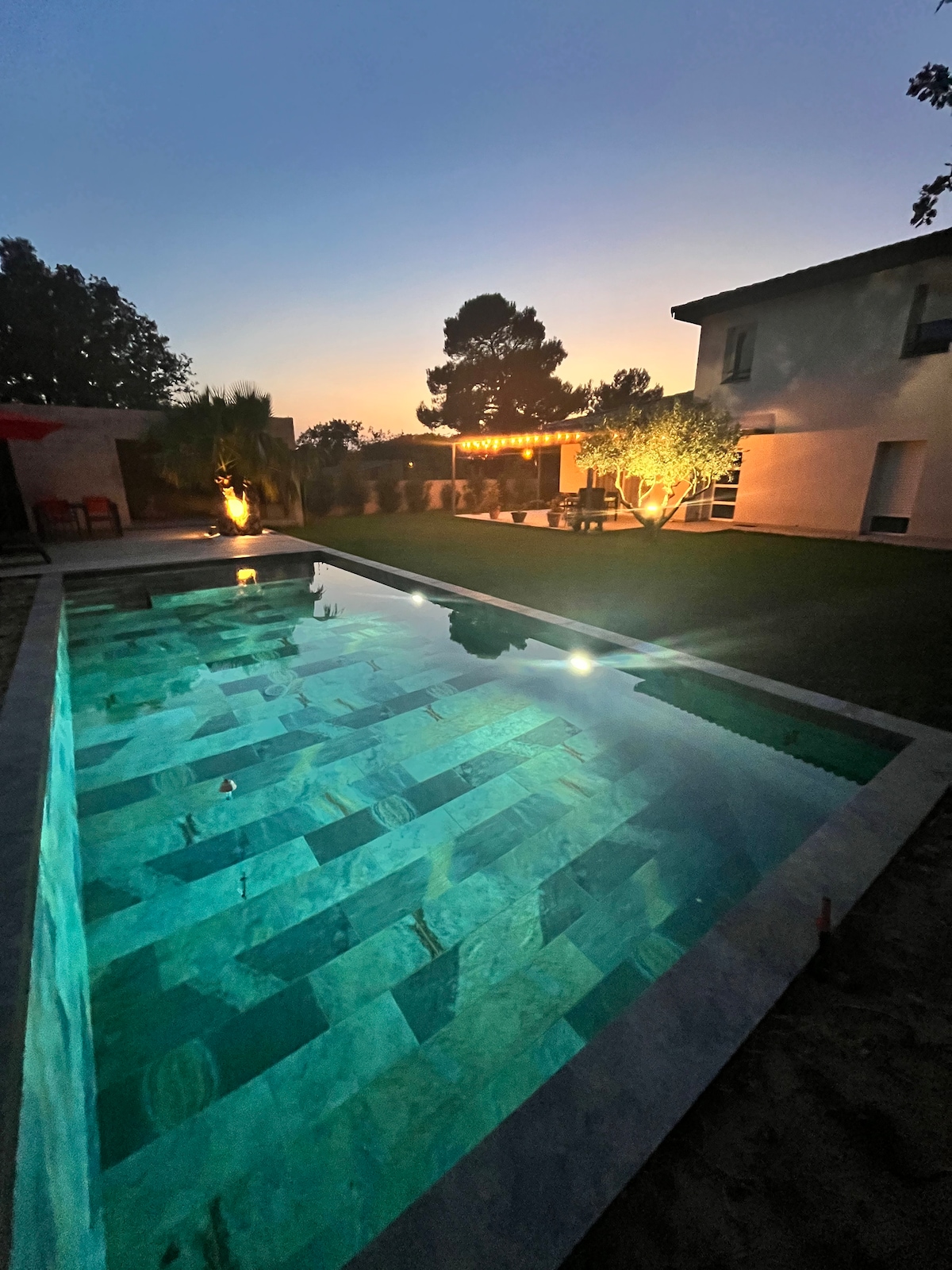A swimming pool features illuminated water reflecting the evening sky. Surrounding the pool are lush gardens and a patio area with ambient lighting. The villa's exterior is visible in the background, providing a serene backdrop for outdoor relaxation.