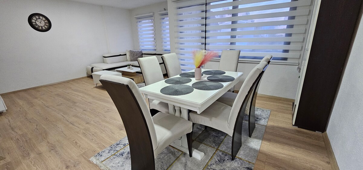 A bright dining area features a white table with six chairs set on a patterned rug. Large windows with striped shades allow natural light to fill the space, complemented by a cozy seating area in the background, creating a welcoming atmosphere.